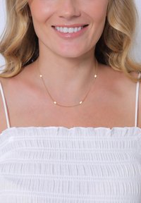 Collier en chaîne dorée présentant de petites perles ovales et rondes alternées. Design léger et élégant, porté avec un haut blanc texturé.