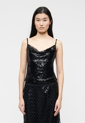 Noor naine, kellel on pikad tumedad juuksed, seljas must helkiv siiditopp ja sobiv must helkiv siidiseelik, seisab ühevärvilise tausta ees.