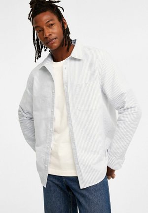 Chemise rayée bleu clair avec un devant boutonné, une poche sur la poitrine et des manches longues, assortie d'un t-shirt blanc à col rond et d'un jean foncé.