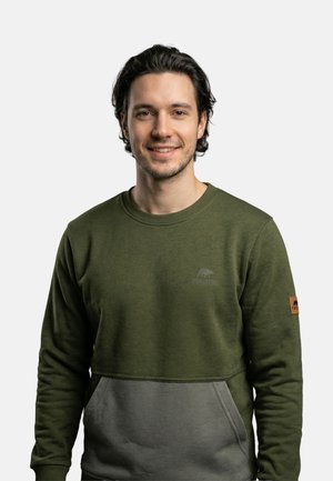 Junger Mann mit dunklen Haaren, der lächelt und ein zweifarbiges Sweatshirt in Olivgrün und Grau mit einer Fronttasche trägt, steht vor einem einfarbigen hellen Hintergrund.