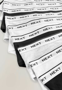 Tricouri din bumbac întinse în culorile negru și alb, cu branding-ul "NEXT" pe manșete, aranjate plat într-o suprapunere.