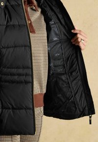 Mustade pufvest, millel on tikitud tekstuur, lahti tõmmatuna, et näidata mustriga sisemist voodrit, kantud pruuni houndstooth särgi ja pükste peal.