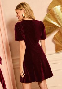 Burgundy bársonydressz rövid puffos ujjal és igazított felsőrésszel. Hátul ipari cipzárral záródik, és sima textúrájú, bővülő szoknyája van.