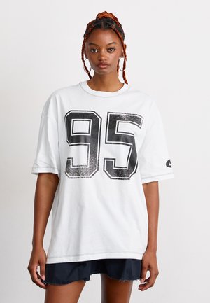Femme avec des cheveux tressés portant de grandes boucles d'oreilles en forme de cercle, un t-shirt oversize blanc avec "95" et un short noir, le tout sur fond uni.