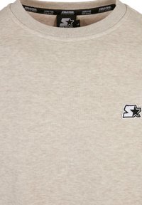 Sudadera beige de cuello redondo hecha de tela texturizada, con un pequeño logotipo de estrella negra bordado en el área del lado izquierdo del pecho.