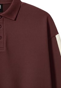 Polo à manches longues marron foncé avec trois boutons et une bande blanche le long de la manche extérieure.