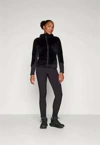 Must karvane tõmblukuga fliiskindel jakk koos tekstureeritud pinnaga, ühes mustadelegaanide ja mustade treenimissussidega, neutraalse tausta ees.