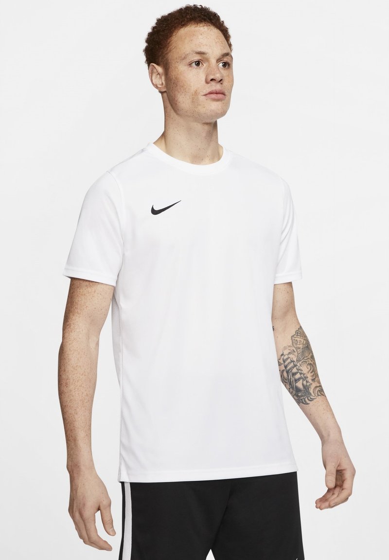 Witte Nike sport t-shirt met korte mouwen, ronde halslijn en zwart logo op de linkerkant van de borst; gedragen met zwarte shorts en zichtbare armtatoeage.