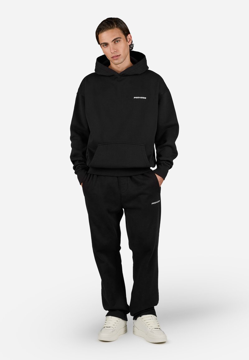 Conjunto de sudadera con capucha negra y pantalones de chándal que cuenta con un bolsillo frontal y texto en logo blanco. Material suave, corte relajado y puños acanalados. Zapatillas blancas.