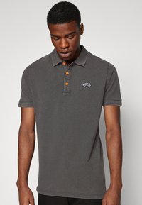 Polo de color gris oscuro hecho de algodón con un cuello clásico, que presenta tres botones naranjas y un pequeño logo negro en el lado izquierdo del pecho.