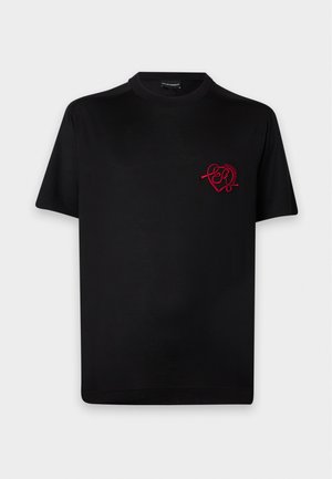 Mand står med den ene hånd i lommen iført sort T-shirt med rødt hjerteprint og lyseblå vidde jeans mod hvid baggrund.