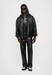 Homme portant une veste et un pantalon en cuir noir sur un sweat à capuche noir avec un design graphique, des lunettes de soleil sombres et des mocassins noirs et blancs.