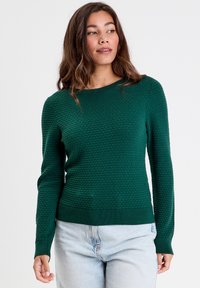 Pull en tricot vert avec un motif en nid d'abeille texturé, col rond et poignets côtelés. Associé à un pantalon en denim clair.
