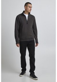 Felpa grigia con zip e collo alto, polsini e orlo a costine, abbinata a jeans neri e sneakers scure. Design semplice e pulito.