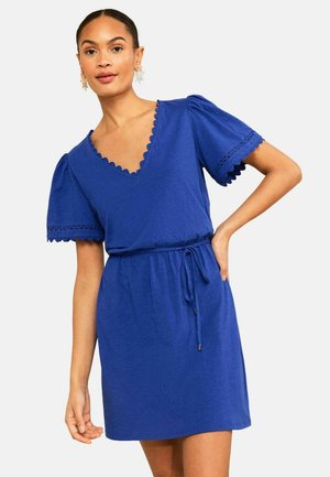 Frau trägt ein knielanges blaues Kleid mit gewelltem V-Ausschnitt, Puffärmeln, Lochstickerei und einem gebundenen Taillengürtel und posiert mit der Hand in der Hüfte.