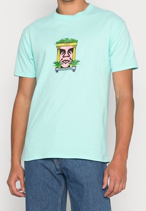 Mand iført en lyseblå T-shirt med abstrakt ansigt indrammet af gule søjler og grønne planter, kombineret med blå jeans.