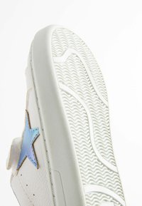 Sneaker bianco con suola in gomma testurizzata, caratterizzato da un accento blu a forma di stella e materiale in pelle liscia. La vista laterale mette in evidenza i dettagli della cucitura.