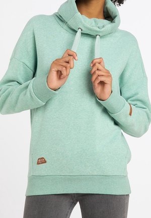 Personne portant un sweat à capuche vert menthe tenant les cordons, visible du cou jusqu’aux hanches, associée à un pantalon gris foncé.