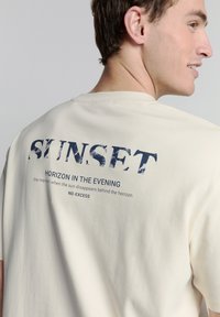 Lichtbeige katoenen T-shirt met een grafische print op de achterkant met blauwe tekst "SUNSET HORIZON IN THE EVENING" en de merknaam.