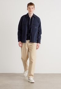 Marineblauwe jas met twee borstzakken, met een opengeslagen kraag, gedragen over een zwarte top. Beige broeken en witte sneakers maken de outfit compleet.