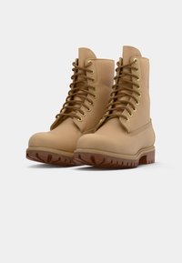 Bottines de travail en cuir tan à lacets avec des semelles en caoutchouc épaisses et des œillets dorés, présentées côte à côte sur fond blanc.