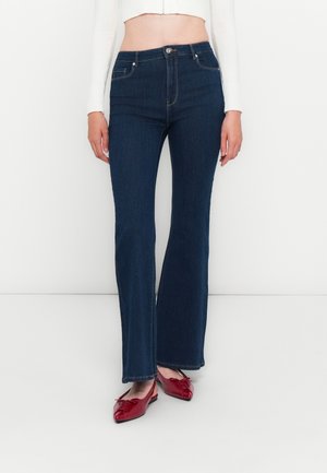 Femme portant un jean évasé bleu foncé, un pull court blanc et des ballerines à talons rouges dans un cadre blanc uni.
