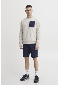 Sudadera beige con un bolsillo azul marino, combinada con pantalones cortos azul marino. El atuendo presenta un ajuste relajado y textura, complementado por zapatillas deportivas oscuras.