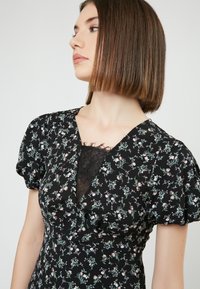 Vestido negro con flores y mangas cortas tipo globo, que presenta un inserto de encaje en el escote. Pequeños patrones de flores en rosa y verde por toda la prenda.