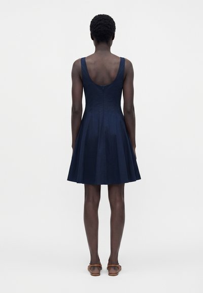 Personne debout de dos, portant une robe bleu marine sans manches et des sandales marron sur un fond clair uni.
