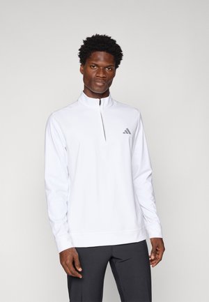 Pull-on athlétique zippé blanc avec des manches longues, fabriqué en tissu lisse. Il présente un petit logo gris sur la poitrine et un design ajusté.