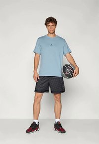 Vīrietis zila T-kreklā, melnos rakstainos īsos šortos un sarkani-melnos sporta apavos tur pāri melnu un pelēku basketbola bumbu uz viendabīga fona.