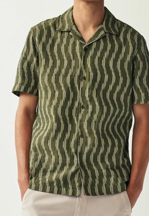 Homme portant une chemise olive à manches courtes avec boutons et un motif zigzag vertical, associée à un pantalon beige clair.