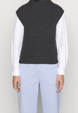 Personne portant un gilet sans manches en maille gris foncé sur une chemise blanche à manches longues et un pantalon bleu clair sur un fond uni.