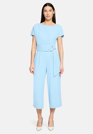 Frau trägt einen hellblauen Jumpsuit mit weitem Bein, kurzen Ärmeln, tailliert mit Gürtel und weißen Pumps mit spitzer Kappe, steht vor einfachem Hintergrund.