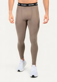 Mann trägt taupefarbene Sportleggings mit schwarzem elastischem Bund mit der Aufschrift "SMILODOX" und weiße Sportschuhe vor neutralem Hintergrund.