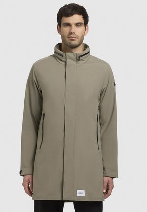 Trench de lluvia largo en verde oliva con un alto cuello, cierre frontal con cremallera y dos bolsillos laterales con cremalleras negras. Fabricado en material suave e impermeable.