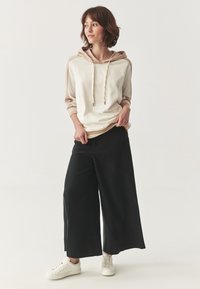 Conjunto informal que incluye una sudadera crema y beige con cordones y mangas largas, combinada con pantalones anchos negros y zapatillas blancas.