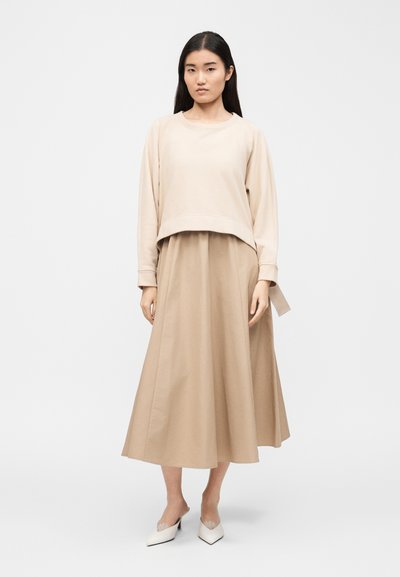 Γυναίκα που στέκεται, ντυμένη με μπεζ μακρυμάνικη μπλούζα, ταιριαστό μεσαίο φόρεμα και λευκά παπούτσια με μυτερή μύτη, μπροστά σε απλό φόντο.