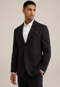 Schwarzer maßgeschneiderter Blazer mit Reverskragen, einem Knopfverschluss und zwei vorderen Taschen. Weißes Hemd darunter mit Knopfdetails.