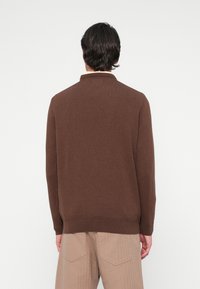 Sötét hajú férfi barna hosszú ujjú pulóverben és bézs nadrágban áll, háttal a kamerának, fehér háttér előtt.