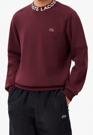 Man wearing a burgundy Lacoste sweatshirt with logo on chest and collar, hands in pockets of black textured pants with Lacoste logo.