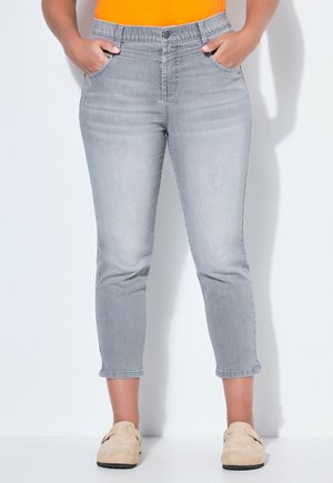 Frau steht mit Händen in den Taschen, trägt hellgraue, gekürzte Jeans, ein orangenes Oberteil und beige Slipper vor weißem Hintergrund.