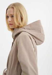 Beige wasserdichte Jacke mit Kapuze, Frontreißverschluss und Kordelzug in der Taille. Glatte Textur, minimalistisches Design und dezente Akzente.