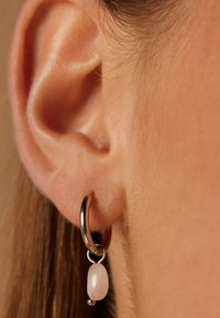 Boucle d'oreille en métal argenté avec un charme en pierre rose détachable. La boucle d'oreille présente une forme lisse et arrondie avec une finitions subtilement brillante.