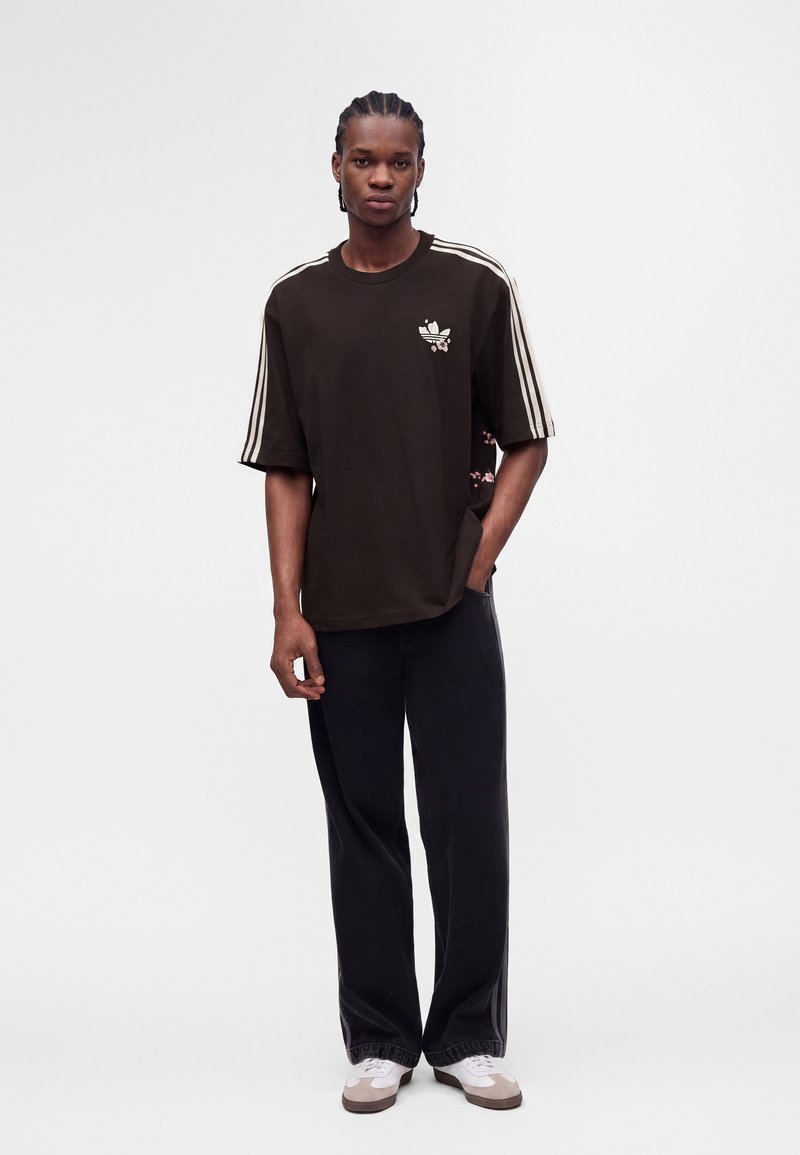 Νέος άνδρας με πλεξούδες, φορώντας μαύρο t-shirt Adidas με λευκές ρίγες, μαύρο παντελόνι και λευκά αθλητικά παπούτσια, στέκεται με το ένα χέρι στην τσέπη.