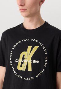 Melna kokvilnas T-kreklu ar lielu grafiku, kurā redzams "Calvin Klein" baltā un dzeltenā krāsā, apaļais dizains un īsām piedurknēm.