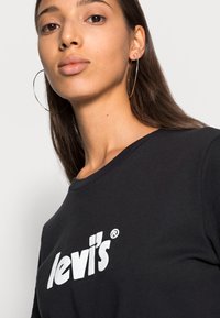 Camiseta de algodón negra con el logo blanco de "levi's", con cuello redondo y mangas cortas, llevada por una persona con cabello largo.