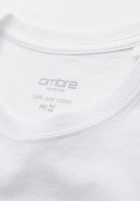 Gros plan sur l'encolure d'un T-shirt en coton blanc montrant l'étiquette de la marque "ombre", taille moyenne, et tissu 100 % coton pur.