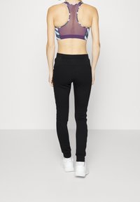 Schwarze, enganliegende Jogginghosen mit einem hohen Bund, kombiniert mit einem lila und gemusterten Sport-BH. Das Outfit hat ein Racerback-Design und besteht aus elastischem Material.