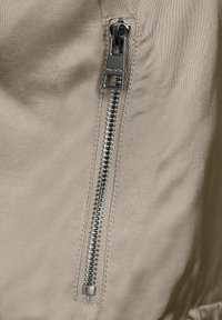 Un tissu beige avec une texture lisse présente une fermeture éclair métallisée argentée sur le côté, mise en valeur par des coutures contrastées.